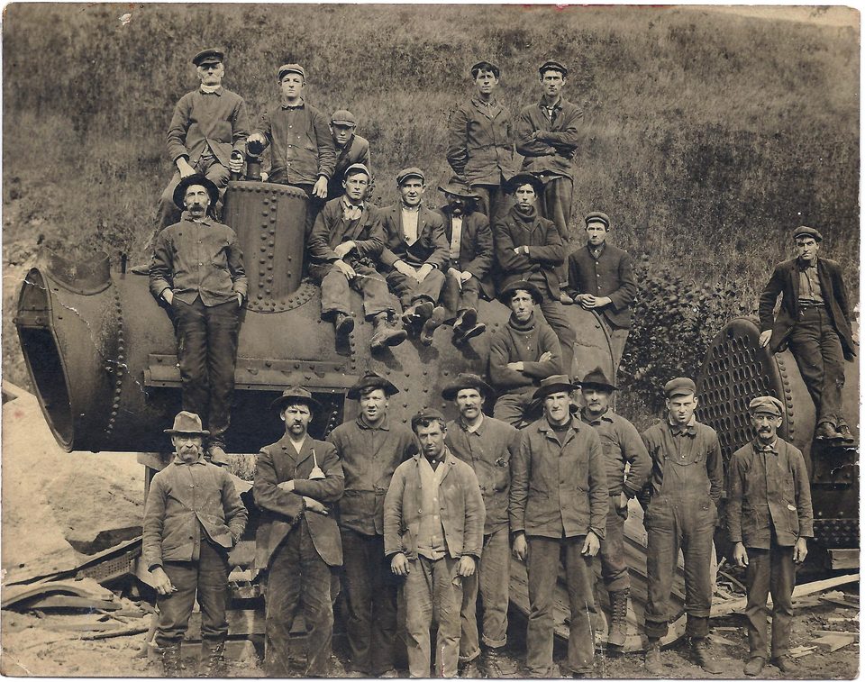 Sepia group photograph of more than twenty men in work clothes, caps and hats, sitting and standing on and in front of a large riveted boiler on a grassy bank.