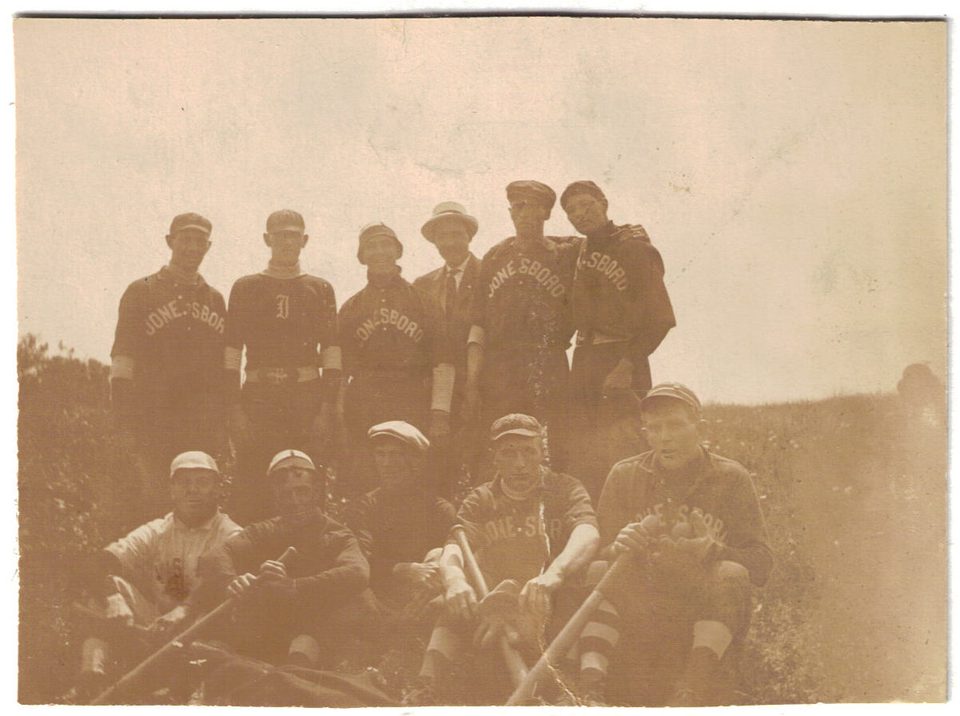 Faded sepia photograph of a baseball team on a hillside, most players in dark shirts lettered JONESBORO, with a man in a suit and straw hat among them.