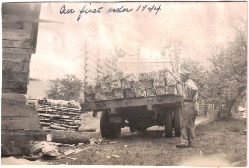 Photograph of a man in a hat beside a flatbed truck stacked with bundles of sawn lumber, with a wooden building at the left; handwritten across the top: Our first order 1944.