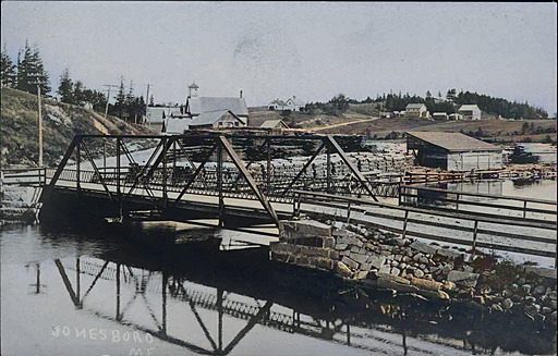 Colourized postcard of a steel truss bridge over still water, its reflection below, with houses and mill buildings on the far bank.
