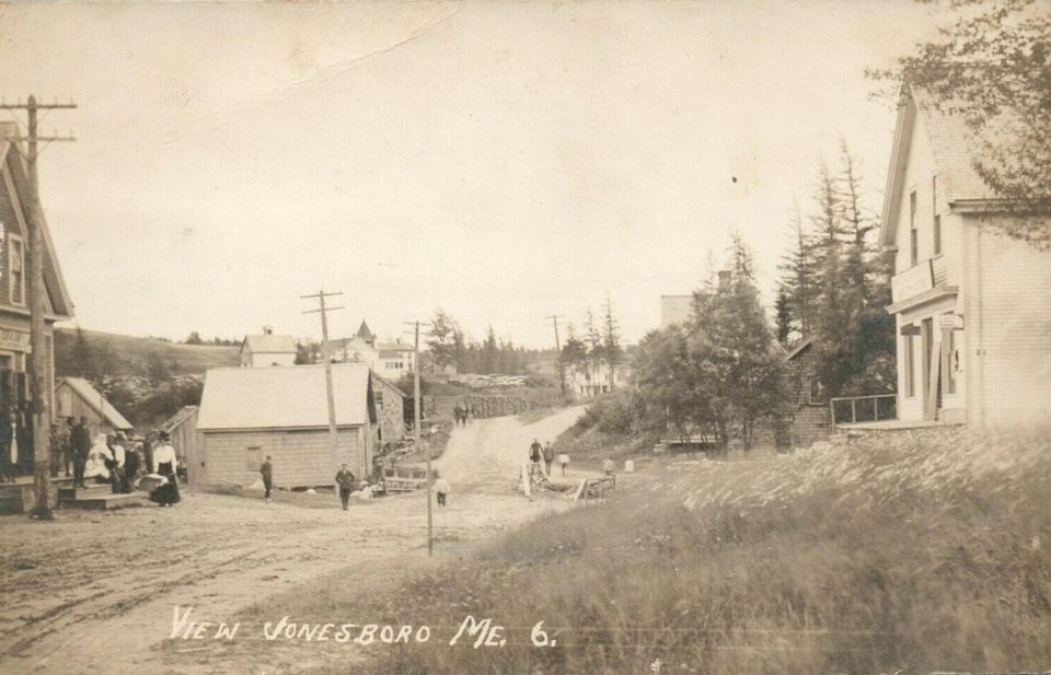 Sepia real-photo postcard of an unpaved village street, with people gathered outside a building on the left, telegraph poles, and white clapboard houses on the right.