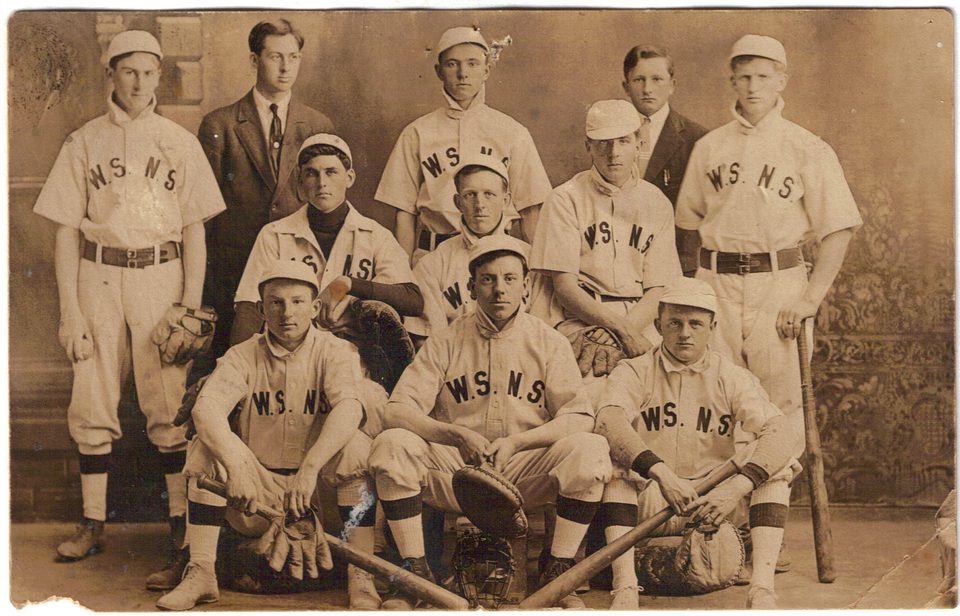 Sepia studio portrait of a baseball team in light uniforms lettered W.S.N.S., with gloves, a catcher's mitt and bats, and two young men in suits in the back row.
